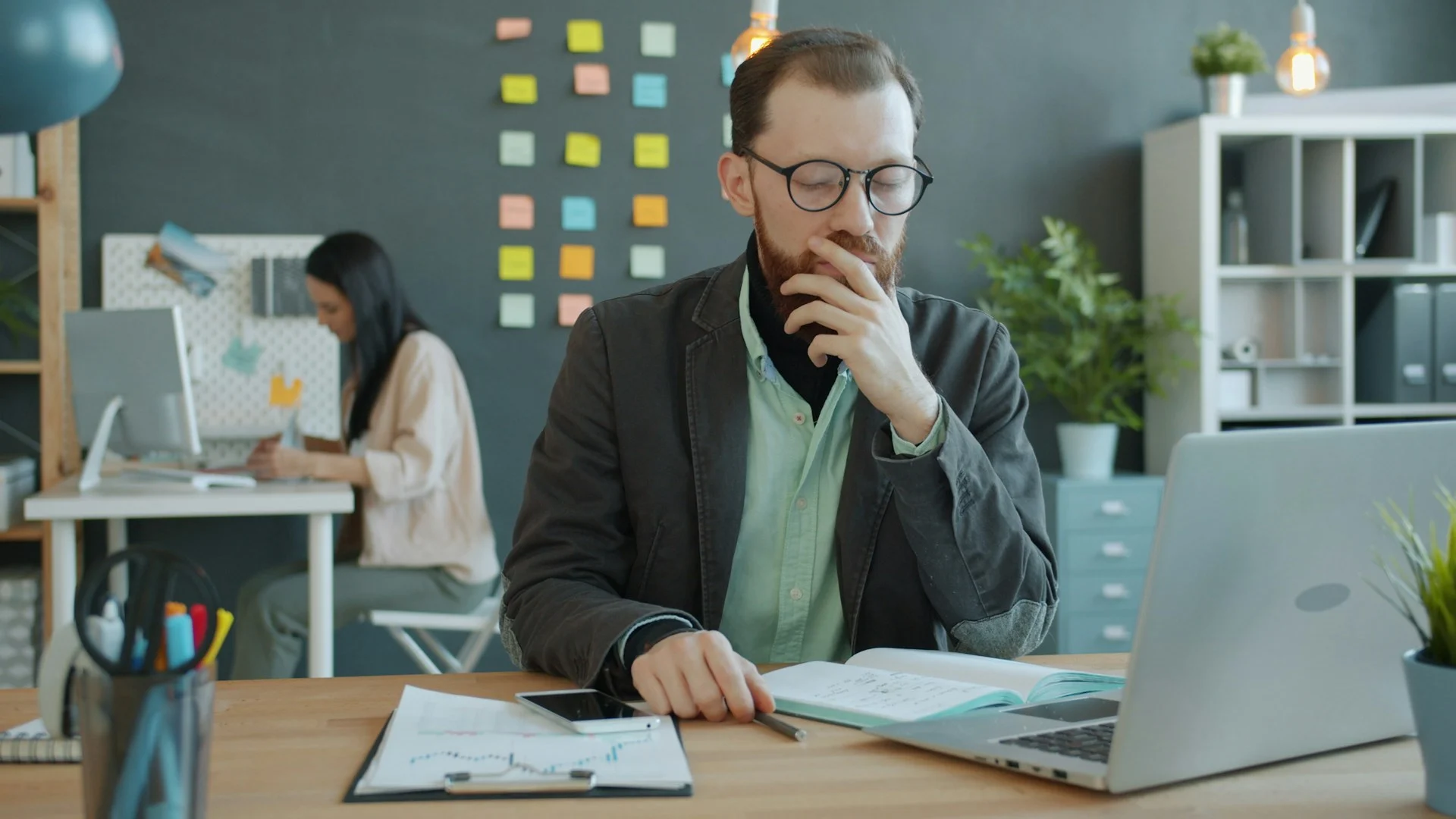 Man thinking at a desk with a laptop in a busy office